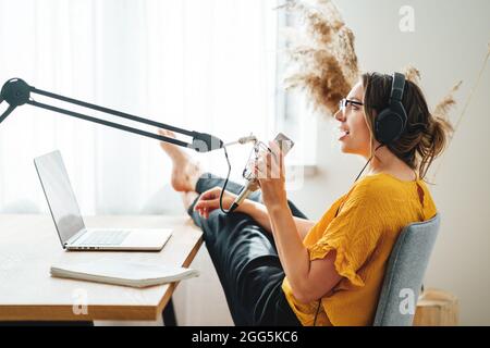 Donna podcaster che trasmette e trasmette il suo podcast da casa studio. Donna che registra podcast da casa e parla in un microfono. Foto Stock