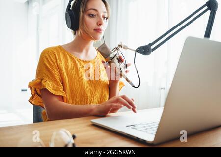 Podcaster femminile che registra il suo podcast utilizzando il microfono e il portatile presso il suo studio di trasmissione a casa. Donna in streaming dal vivo da casa studio radio Foto Stock