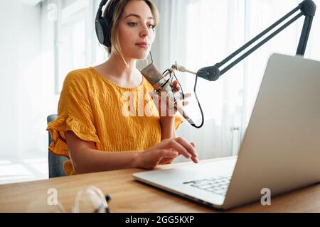 Giovane ospite radiofonico femminile che registra e trasmette il suo podcast da studio fatto in casa. Donna podcaster parlare nel microfono sul tavolo Foto Stock