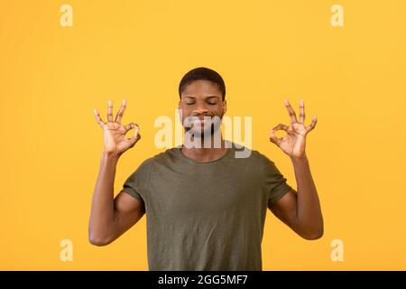 Concetto di sollievo dallo stress. Giovane uomo nero meditando con occhi chiusi, mantenendo la calma su sfondo giallo studio Foto Stock