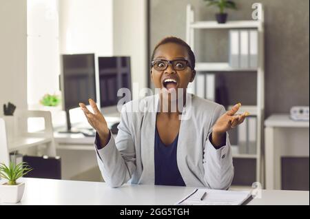 Felice sorprende donna nera seduta alla scrivania dell'ufficio, guardando la macchina fotografica e applaudendo Foto Stock