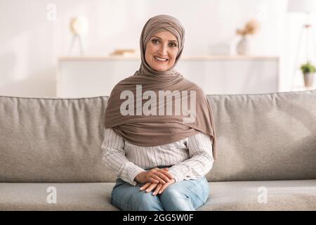 Donna islamica senior positiva indossando Beige Hijab seduta a casa Foto Stock