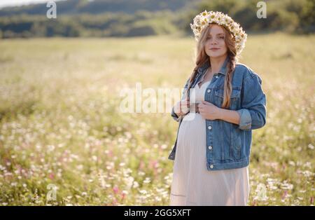 Bella donna incinta in un abito, giacca in denim e una corona di margherite rilassante fuori nel parco. Gravidanza sana e concetto di viaggio. Foto Stock