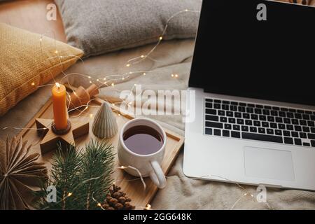 Computer portatile su letto morbido con tazza di tè, luci di natale, stelle, pino, candela e cuscini in camera scandinava. Freelance e blog. Lavoro in remoto e online Foto Stock