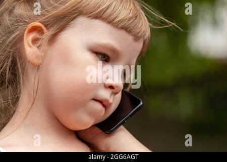 Bambino giovane, bambina profonda nel pensiero parlare sul telefono cellulare, smartphone. Ritratto naturale, espressione neutra, faccia closeup estremo. Telecomm Foto Stock