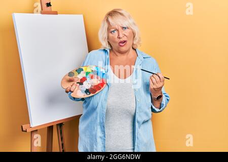 Donna bionda di mezza età in piedi disegno con tavolozza da pittore cavalletto in piedi in faccia shock, guardando scettico e sarcastico, sorpreso con bocca aperta Foto Stock