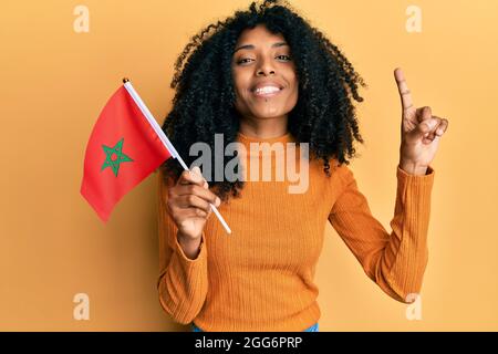 Donna afroamericana con capelli afroamericani che reggono la bandiera del marocco sorridendo con un'idea o una domanda puntando il dito con il volto felice, numero uno Foto Stock