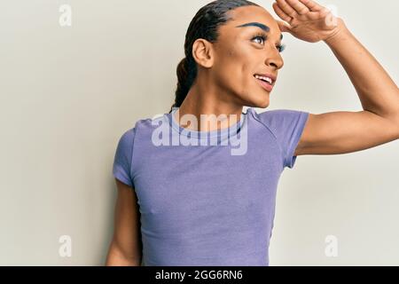 Uomo ispanico che indossa make up e lunghi capelli che indossa casual t shirt molto felice e sorridente guardando lontano con mano sulla testa. Concetto di ricerca. Foto Stock