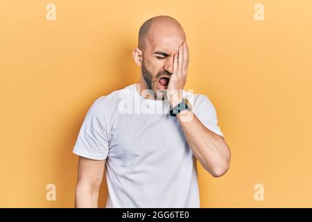 Giovane uomo calvo che indossa casual maglietta bianca che sbadigia stanco coprendo metà faccia, occhio e bocca con mano. Il viso fa male nel dolore. Foto Stock