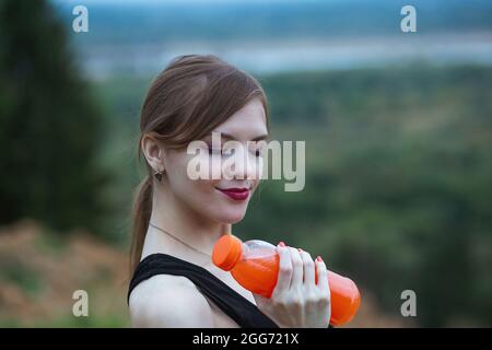 Donna che indossa un abbigliamento sportivo che si idrata bevendo succo d'arancia vivace durante l'allenamento. Sera o mattina nella natura. Foto Stock