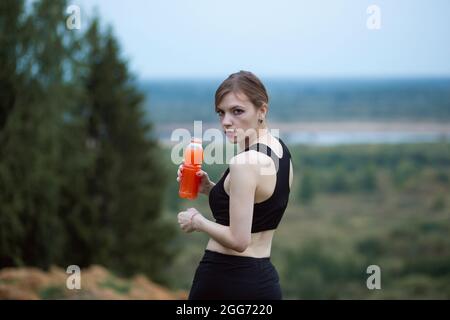 Donna che indossa un abbigliamento sportivo che si idrata bevendo succo d'arancia vivace durante l'allenamento. Sera o mattina nella natura. Foto Stock