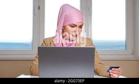 Giovane donna musulmana in un foulard rosa fa acquisti online su un portatile che tiene una carta di banca nelle mani Foto Stock