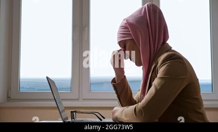 Donna in hijab rosa fa una pausa e massaggia il ponte del naso mentre si lavora su un computer portatile Foto Stock