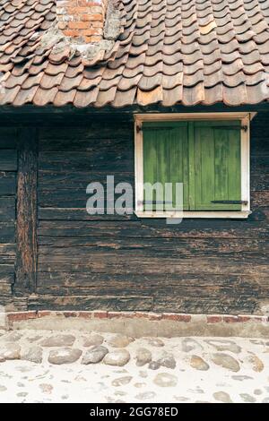 Un vecchio muro in legno con persiane chiuse verdi e tegole rosse, sul lato di un frammento di strada acciottolato di pietra del muro in legno o Foto Stock