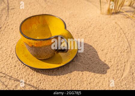 Una tazza di argilla con glassa di vetro giallo e una piattina corrispondente è posta nella sabbia della spiaggia. Viste oblique della tazza con un piatto. Foto Stock