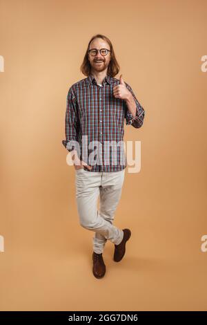 Zenzero bearded uomo che indossa occhiali sorridenti e mostrando pollice su isolato su sfondo beige Foto Stock