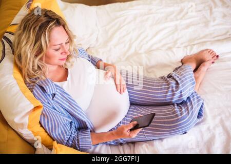 Donna incinta in pigiama blu a strisce che guarda in smartphone in camera da letto la mattina dopo il risveglio. Donna in attesa di un bambino nel nono mese Foto Stock