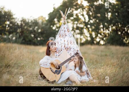 Affascinante giovane donna boho hippie che suona la chitarra per la sua bambina carina all'aperto in natura la sera di sole, seduto di fronte a wigwam tenda e cantare. Tradizioni familiari musicali Foto Stock