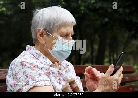 Donna anziana in maschera protettiva seduta con smartphone su una panca in parco. Sicurezza durante la pandemia di coronavirus, vita in pensione Foto Stock