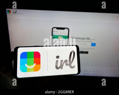 Persona che tiene il cellpohone con il logo del social network degli Stati Uniti IRL (Get Together Inc.) sullo schermo di fronte al sito web aziendale. Mettere a fuoco sul display del telefono. Foto Stock