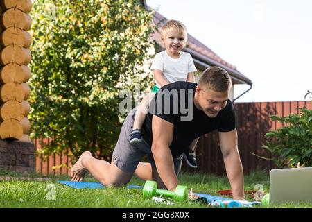 Il giovane uomo con bambino entra per praticare sport in giardino. Lo sportivo con capelli biondi si allena, facendo dei push-up con suo figlio sulla schiena sul prato Foto Stock