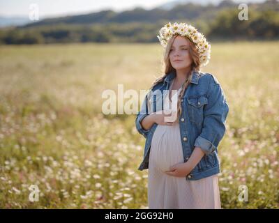 Bella donna incinta in un abito, giacca in denim e una corona di margherite rilassante fuori nel parco. Gravidanza sana e concetto di viaggio. Foto Stock