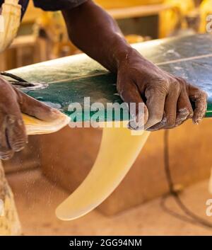 Uno Shaper da surf di un artigiano africano che lavora in un'officina di riparazione Con carta vetrata Foto Stock