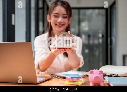 Immobiliare donna mani tenuta casa modello presentare al cliente con Piggy banca su scrivania. Risparmio di denaro vendere o acquistare concetto di casa Foto Stock