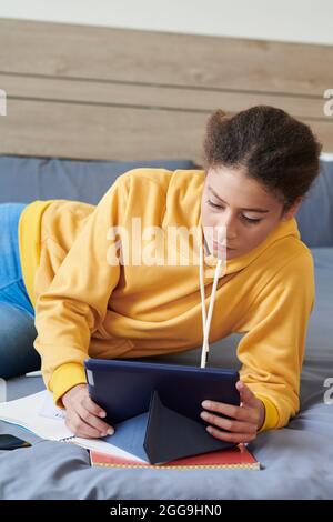 Ragazza seria adolescente in felpa gialla che si trova sul letto nella sua stanza e guardando video educativo Foto Stock