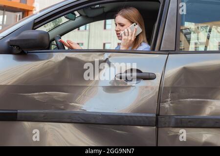 Vista laterale di donna sconvolto con capelli biondi seduta in schiantato auto e telefono di conversazione, chiamata all'agente di assicurazione o al marito, ha problemi con auto d Foto Stock