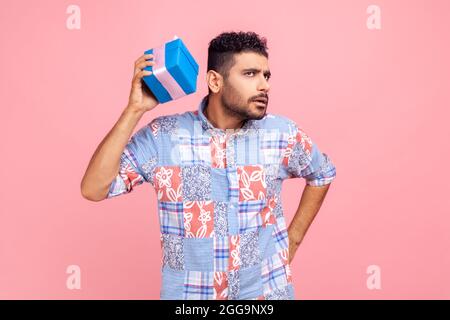 Ritratto di curioso uomo bearded che indossa la camicia di stile casual blu che tiene e scuotendo avvolto scatola presente, essendo interessato che cosa all'interno. Monolocali al coperto Foto Stock