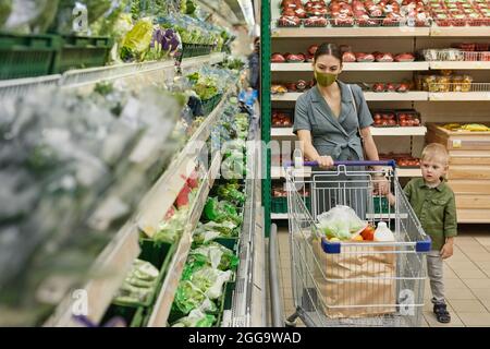 Donna giovane Caucasain incinta in maschera di panno spingendo carrello e facendo acquisti con figlio nel reparto di verdure del supermercato Foto Stock