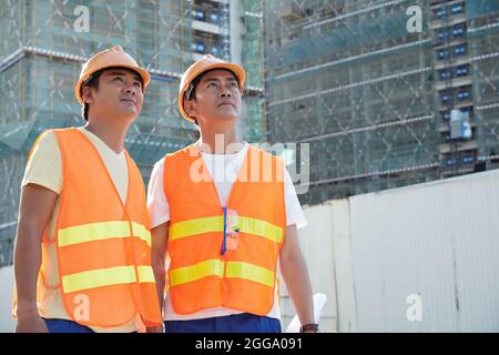 Team di costruttori in giubbotti arancioni e con cappelli che guardavano alti edifici di un nuovo hotel o centro affari Foto Stock