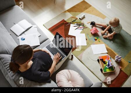 Sopra il viiew della madre incinta occupata usando il laptop mentre suo figlio gioca con i giocattoli sul tappeto nel soggiorno Foto Stock
