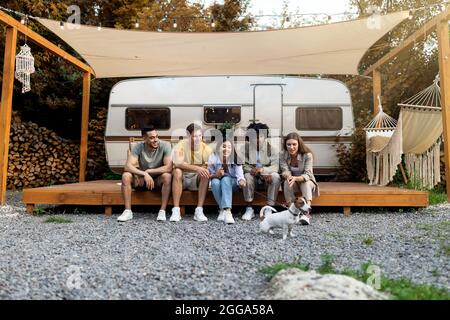 Gruppo di giovani allegri persone diverse con il loro carino cane seduto vicino a casa, avendo divertente viaggio in campeggio in autunno Foto Stock