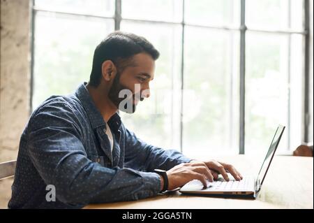 Positivo ragazzo intelligente indiano freelancer che lavora su laptop in ufficio a casa, websurfing, multirazziale uomo dipendente orientale utilizzando il computer per il lavoro remoto, il testo e la digitazione, il programmatore indù è la codifica Foto Stock