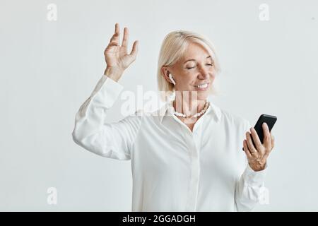 Ritratto in vita di donna matura elegante danza con auricolari wireless su sfondo bianco, spazio copia Foto Stock