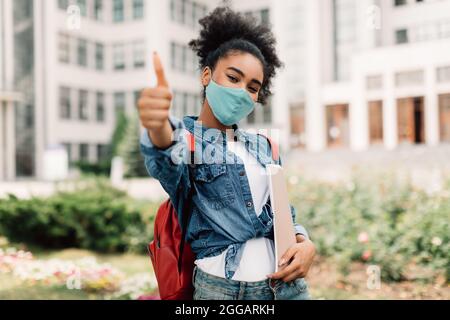 Studentessa nera in maschera facciale gesturando i thumbs-up in posa all'aperto Foto Stock