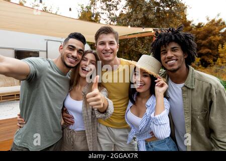 Diversi millennial che prendono selfie insieme vicino a camper van, sorridendo alla macchina fotografica, trascorrendo del tempo insieme in campeggio Foto Stock