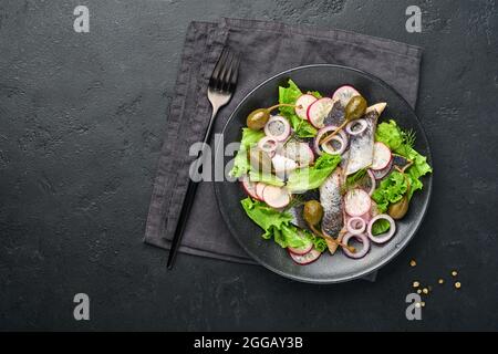 Aringhe salate con spezie, capperi, erbe e insalata di cipolla su piatto nero su sfondo scuro con spazio copia. Pesce marinato a fette. Cibo con u sano Foto Stock