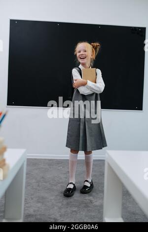 Una scolaretta che abbraccia un libro di testo in piedi di fronte a una tavola da ballo in una classe. Foto Stock