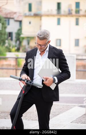 Bel giovane uomo d'affari con computer portatile, in abito formale e su scooter elettrico, sta verificando l'inizio e si affretta a andare al lavoro Foto Stock