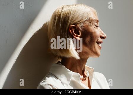 Ritratto minimale della vista laterale di donna matura elegante illuminata dalla luce del sole contro la parete bianca, spazio copia Foto Stock