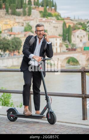 Uomo d'affari con abito elegante in piedi sullo scooter elettrico mentre parla al telefono. Imprenditore che guida lo scooter e risponde al telefono Foto Stock