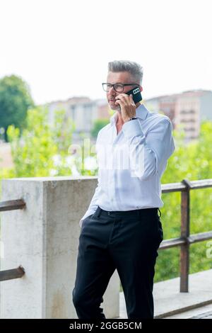 L'uomo d'affari di buon umore sta parlando sul cellulare sul suo senso all'ufficio. Bel manager allegro in tuta formale a piedi godendo di arrivare al lavoro Foto Stock