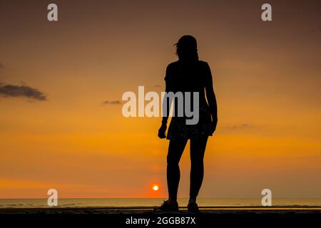 Silhouette posteriore di una giovane donna isolata, in berretto sportivo, guardando al mare al tramonto su una spiaggia del Regno Unito, vacanza estiva staycation. Foto Stock