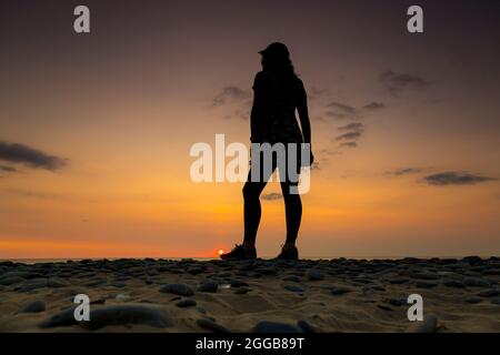 Vista posteriore della giovane donna in berretto sportivo guardando verso il mare e il cielo al tramonto al crepuscolo su una spiaggia del Regno Unito, vacanza estiva staycation. Foto Stock