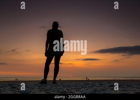 Vista posteriore della giovane donna in berretto sportivo guardando verso il mare e il cielo al tramonto al crepuscolo su una spiaggia del Regno Unito, vacanza estiva staycation. Foto Stock