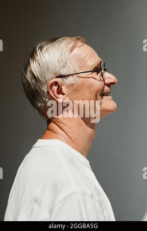 Ritratto con vista laterale minimale di uomo anziano sorridente illuminato dalla luce del sole contro il muro grigio Foto Stock