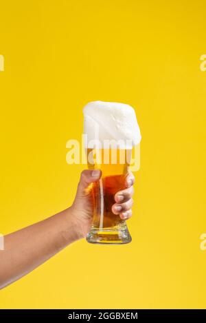 il braccio dell'uomo contiene un bicchiere di vetro con birra all'interno, schiuma bianca e fondo giallo solido, una parte di schiuma cade Foto Stock